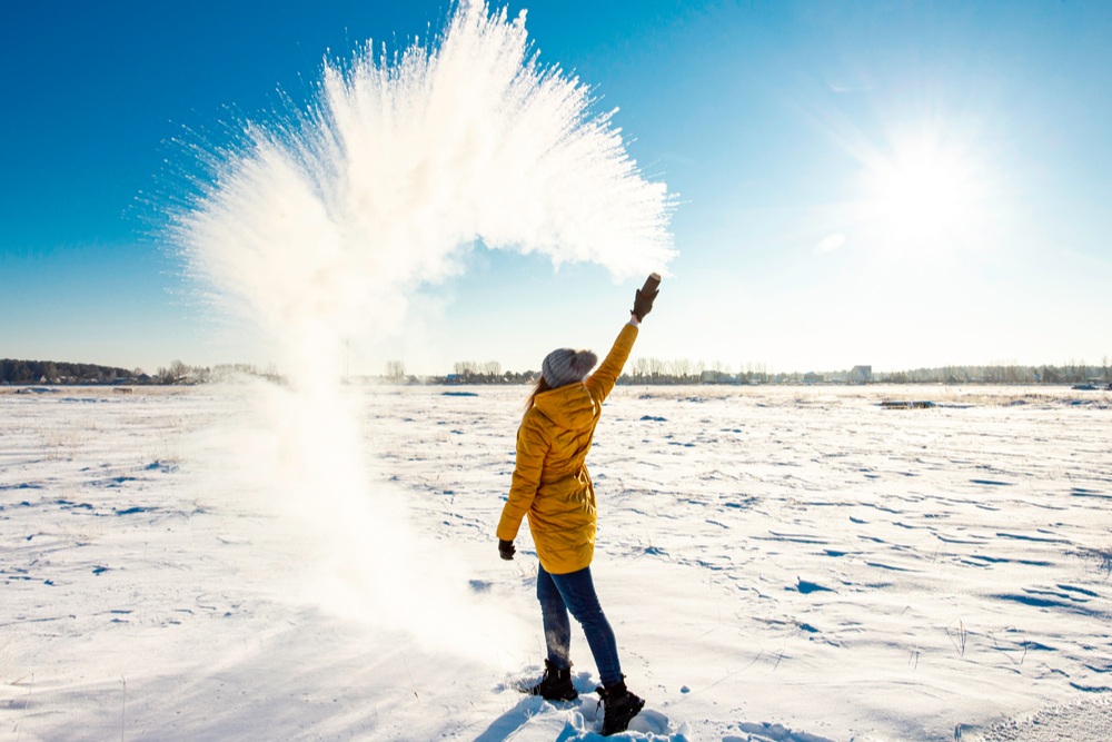 Girl in a bright yellow jacket throws boiling water from a thermos that instantly freezes and turns into an arc of snow